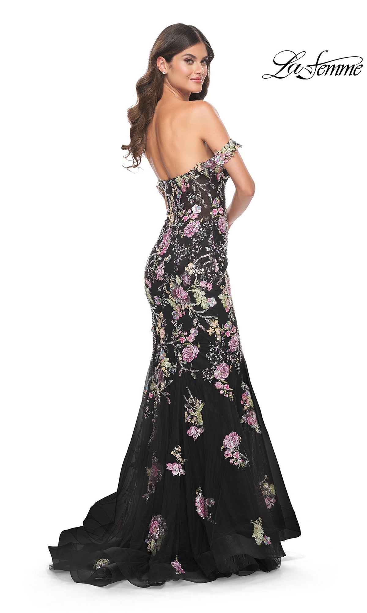 Wearing the La Femme Long Floral Mermaid Prom Dress 32087, a woman poses with her back slightly turned, showcasing a stunning black off-the-shoulder gown with pink and green floral embroidery.